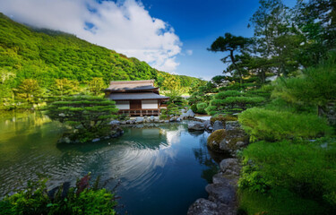 Fototapeta premium A small wooden structure nestled in a lush green garden. The garden features a tranquil pond with a rippling surface.