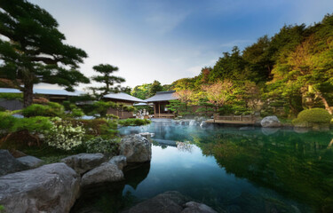 A tranquil Japanese garden features a serene pond with steam rising from its surface, surrounded by lush greenery and traditional wooden structures.