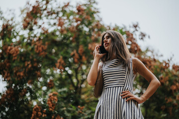 A confident woman stands outdoors in a striped dress, talking on her phone against a backdrop of trees. The mood is empowering and independent.