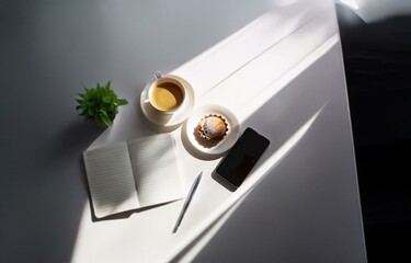 A flatlay of a cup of coffee, a pastry, a notebook, a pen, and a phone on a white surface.