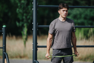 Young man performing calisthenics exercises with a resistance band in an outdoor park environment on a sunny day.