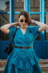 Confident woman in blue dress and sunglasses striking a pose outdoors. Fashionable and stylish female adjusting her hair in a modern setting.