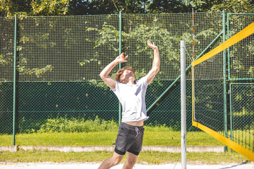 Young man is in mid-air, performing a powerful volleyball jump serve on a sandy court, with his arm raised to strike the ball. The intense action takes place outdoors, surrounded by a green backdrop