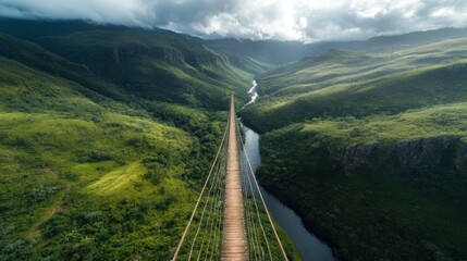 Fototapeta premium A stunning aerial view of a suspension bridge crossing a vibrant green valley, with a river meandering below and mountains receding into the cloudy distance.