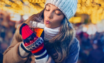 Girl with knitted gloves cooling a hot drink by her breathe under the lights of a winter market