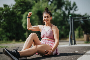 Confident woman in pink sportswear sits on ground at outdoor gym, flexing bicep and smiling.
