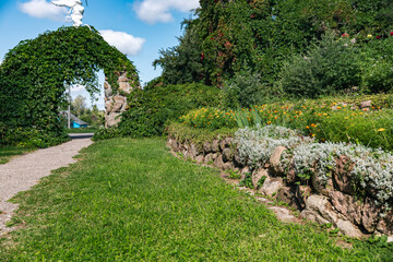 Serene garden scene. A stone archway. The garden is vibrant and well-maintained, with lush green grass, blooming flowers in various shades, and a stone wall.