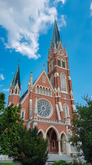 Fototapeta premium Majestic church Herz Jesu Kirche in Graz, Styria, Austria. Landmark against clear blue sky. Intricate architectural details, including pointed spire, ornate windows, clock face. Lush green foliage