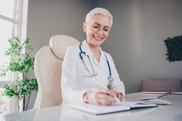 Photo of aged woman qualified doctor pen write notepad wear white coat workplace office indoors