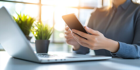 Obraz premium successful businesswoman is using smartphone while sitting at modern desk with laptop and potted plants. bright sunlight creates warm and productive atmosphere