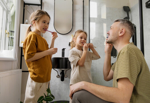 A father teaches his two daughters how to brush their teeth in the bathroom. He is showing them how to hold the toothbrush correctly and how to brush their teeth properly.
