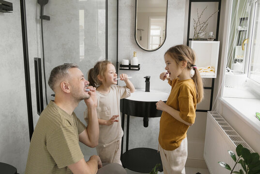 A father is teaching his two daughters how to brush their teeth in the bathroom. They are all smiling and enjoying the experience.
