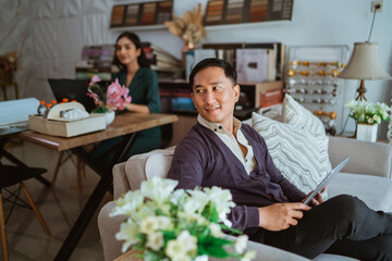 asian male shop owner looking back while sitting on sofa with the digital tablet at his hand inside the interior shop