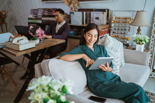 asian beautiful shop owner watching her digital tablet while sitting on sofa with the male shopkeeper at the background inside the interior shop