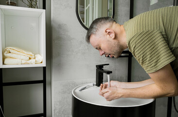 A man is washing his face in a modern bathroom. He is bending over the sink, with water running...