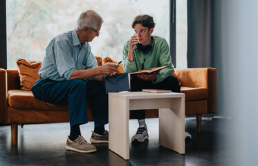 A mature man and a young adult sit across from each other on a couch, engrossed in conversation and...