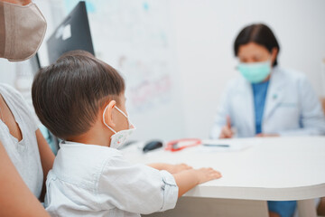 Fototapeta premium A young child wearing a mask sits with their guardian at a doctor's office, consulting with a doctor for medical advice and check-up.