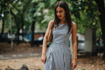 Fototapeta premium A young woman wearing a stylish striped dress enjoys a peaceful walk in a green park. She exudes confidence and contentment amidst the natural summer setting.