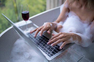 Sexy woman relaxing in a bathtub filled with bubbles also a glass of red wine next to the laptop with speak smartphone is holiday relaxing concept.