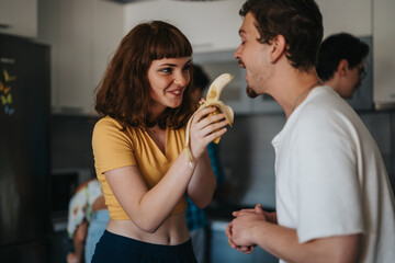 A cheerful group of friends sharing a lighthearted moment in a cozy kitchen, with a woman playfully holding a banana to a smiling friend.
