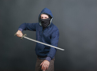 A man wearing a blue hoodie with the hood up and a black face covering, leaving only his eyes visible holds a sword, showcasing a dynamic pose. A person poses with a sword against a dark gray backdrop