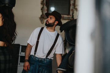 Young man in casual attire wearing suspenders and a hat, standing in a rustic setting with horse equipment in the background.