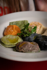 A vibrant plate of sorrentinos and ravioli showcasing different flavors and colors. The dish includes green, orange, black, and white pasta varieties, drizzled with olive oil and garnished with fresh 