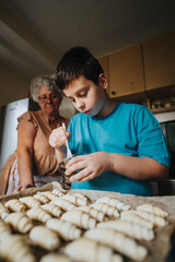 A young boy and his grandmother are in the kitchen baking pastries together, creating a warm family moment. The boy is focused on his task while the grandmother guides him.