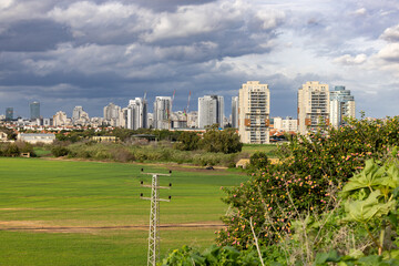 Obraz premium Hod Hasharon, Israel 13 Jan 2024. A panoramic view of the town from a distance. The city skyline is visible with numerous high-rise buildings, surrounded by green fields and a cloudy sky
