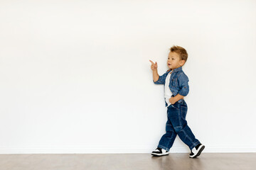 A cheerful toddler strolls with one hand in their pocket and the other pointing, dressed in a trendy denim jacket and jeans while surrounded by a minimalist backdrop. Copy space