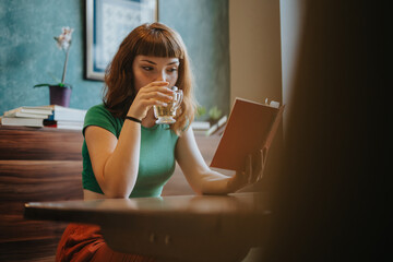 A woman in a green shirt enjoys reading a book while sipping tea in a cozy setting. The image...