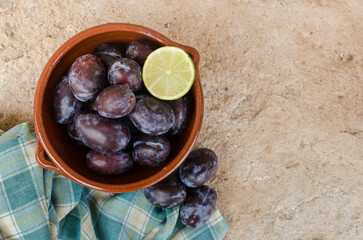 Horizontal close up photography of artistic composition of pottery bowl full of fresh black plums decorated with half lemon and rustic cloth. Fresh ecologic fruit concept. Copy space.