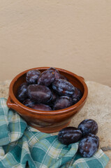Vertical close up photography of artistic still life composition of clay bowl full of fresh black plums decorated with green cloth. Fresh organic fruit concept. Out of focus background. Copyspace.