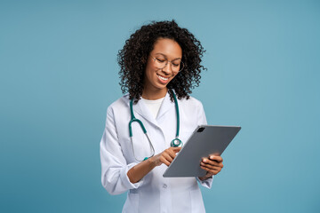 Female doctor using digital tablet, wearing lab coat and stethoscope standing isolated on beige