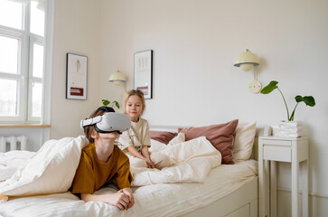 Fototapeta premium Two young girls are in a bedroom, with one wearing a VR headset and looking at the virtual world, while the other girl observes with a curious and entertained expression.