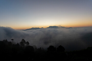 Before sunrise view, painted sky with soft orange light in the horizon. Dense clouds covers the valley and mountains in background above the clouds. Magical morning scene in mountain village in India.