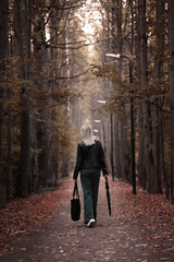 A young blonde girl walks along an autumn alley among the trees. The view from the back.