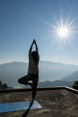 A young woman holds a yoga pose on yoga mat.Sunrise silhouette against layered dusky mountains.The morning light casts sun flares, adding a serene glow to her peaceful practice.Lifestyle in mountains.