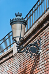 An antique street lamp with a decorative wrought iron bracket and a glass globe. The lamp is mounted on a brick wall and is partially shaded by the shadows of the iron railings above.
