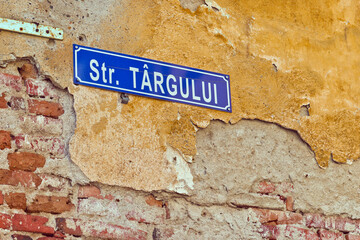 A close-up of a blue street sign for "Str. Târgului" mounted on a weathered brick wall. The wall is covered in peeling paint and has visible cracks and damage. 