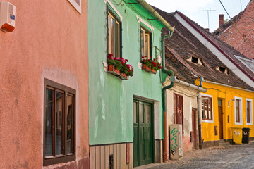A colorful street scene in Sibiu, Romania, featuring a row of historic houses with various pastel hues. The buildings have red-tiled roofs, wooden shutters, and flower boxes on the windows. 