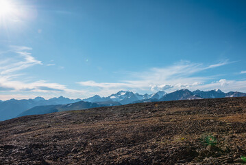 Beautiful big snow-capped mountain top far away in sunny day. Scenic alpine top view from sunlit high stony pass to large rocky snowy mountain peak with glacier in bright sun under clouds in blue sky.