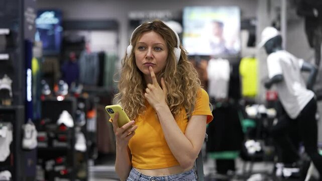 Woman selecting wireless headphones in an electronics store while listening to music through a smartphone app. Engaging in the shopping process, she compares options and makes a decision