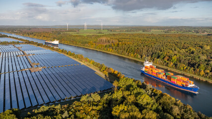 Container ship and car ferry navigate the Kiel Canal alongside vast solar panel fields and wind...