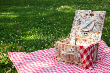 One picnic wicker basket with checkered napkin, tableware and blanket on green grass. Space for text