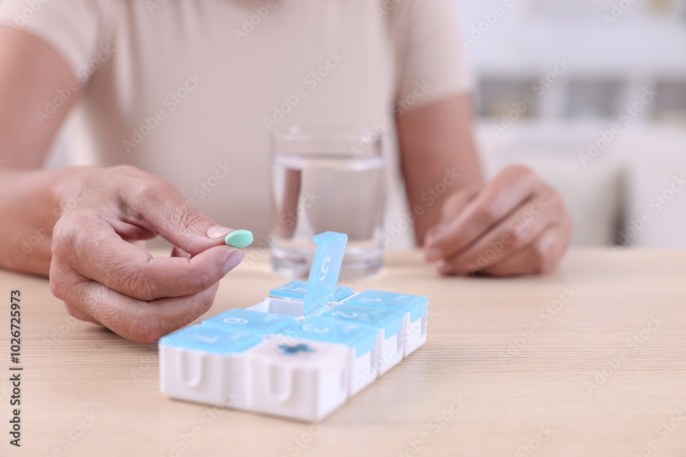 Poster Senior woman taking pill from organizer at table indoors, closeup