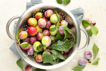 Ripe plums and leaves in colander on light table, flat lay