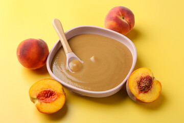Delicious baby food with spoon in bowl and fresh peaches on yellow table