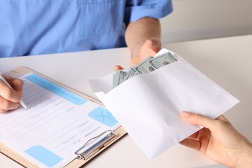 Corruption concept. Woman giving envelope with dollar banknotes to doctor at white table, closeup