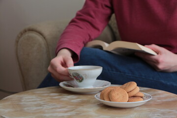 Cookies on a plate and a teacup of tea on a beige marble coffee table. Young woman with book in hand and holding cup for drink tea. Have a good time at home
